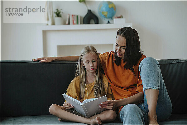 Mother and daughter reading book together on sofa at home