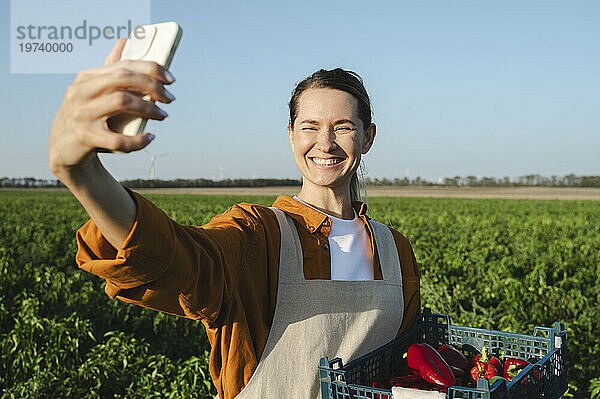 Glücklicher Bauer macht ein Selfie per Smartphone und hält eine Kiste Paprika auf dem Feld