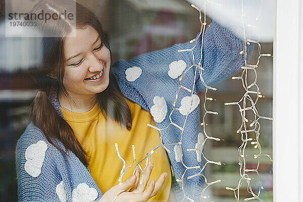 Fröhliches Teenager-Mädchen dekoriert mit Lichterketten  die durch Glas gesehen werden