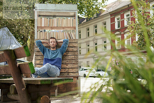 Man with eyes closed leaning on cabinet of books near plants in suburb