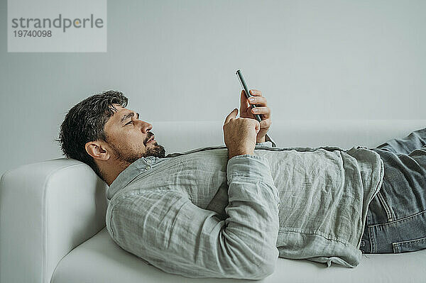 Mann benutzt Smartphone und liegt auf Sofa vor weißem Hintergrund