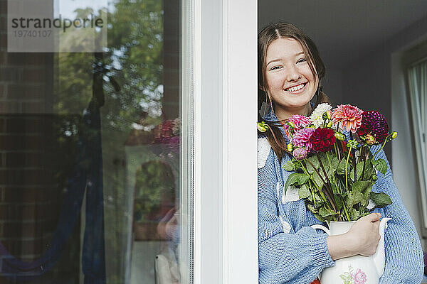 Lächelndes Teenager-Mädchen hält Dahlienblüten in der Hand und lehnt am Fenster