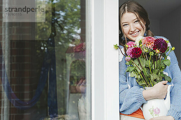 Fröhliches Teenager-Mädchen hält Dahlienblüten in der Hand und lehnt am Fenster