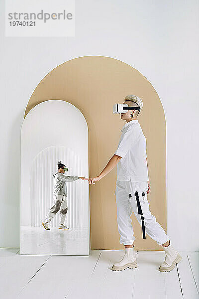Frau mit Virtual-Reality-Headset berührt Spiegelbild eines Mannes im Spiegel an der Wand