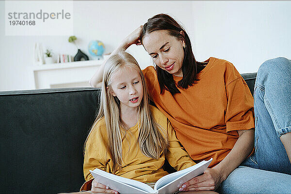 Girl reading book to mother on sofa in living room at home