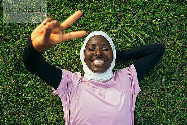 Lächelnde Frau liegt auf Gras und gestikuliert mit einem Friedenszeichen