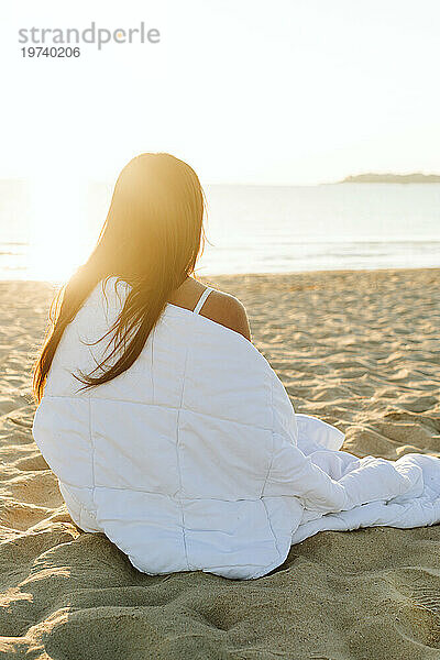 Frau sitzt in Decke gehüllt am Strand an einem sonnigen Tag