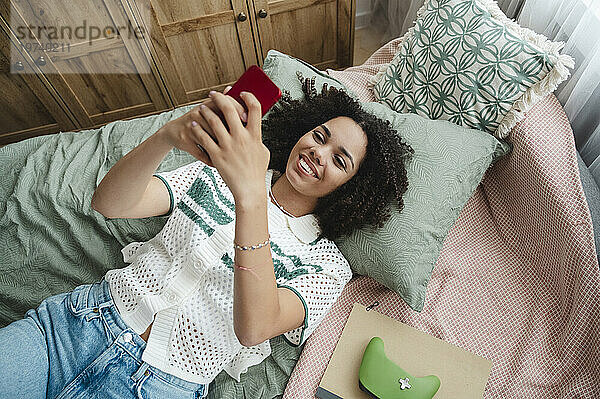 Glückliches Mädchen  das auf dem Bett liegt und zu Hause sein Smartphone benutzt