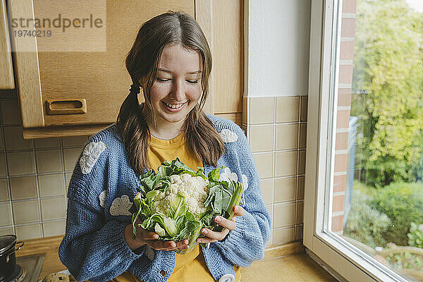 Lächelndes Teenager-Mädchen hält Blumenkohl am Fenster zu Hause