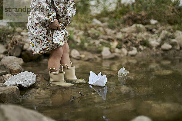 Girl wearing rubber boots and playing with paper boat in water puddle