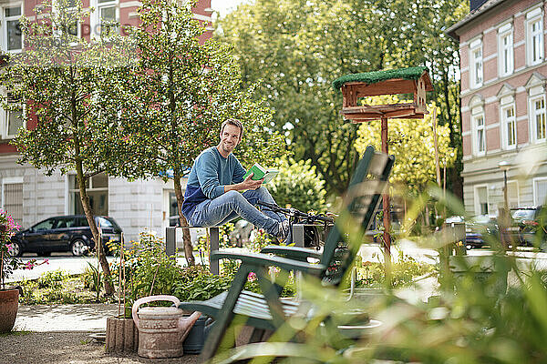Glücklicher Mann hält ein Buch in der Hand und sitzt auf einer Metallstruktur im Garten
