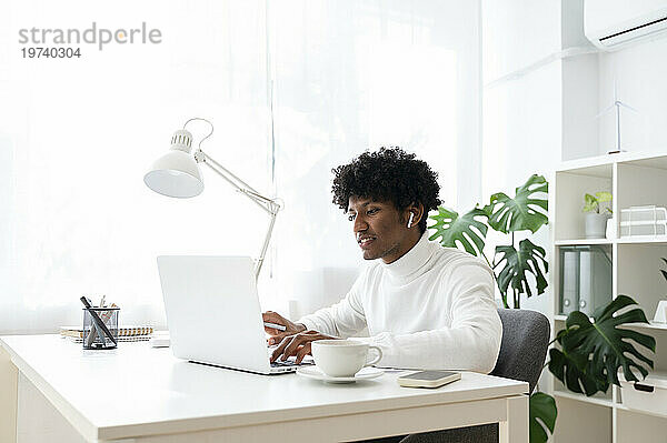 Konzentrierter Geschäftsmann mit kabellosen In-Ear-Kopfhörern  der am Schreibtisch am Laptop arbeitet