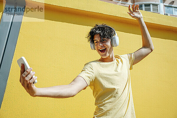 Lächelnder Mann mit kabellosen Kopfhörern macht ein Selfie vor einer gelben Wand