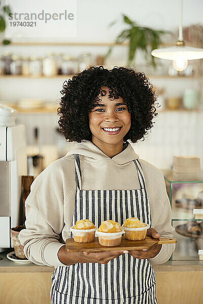 Fröhlicher Barista hält Cupcakes auf einem Schneidebrett im Café