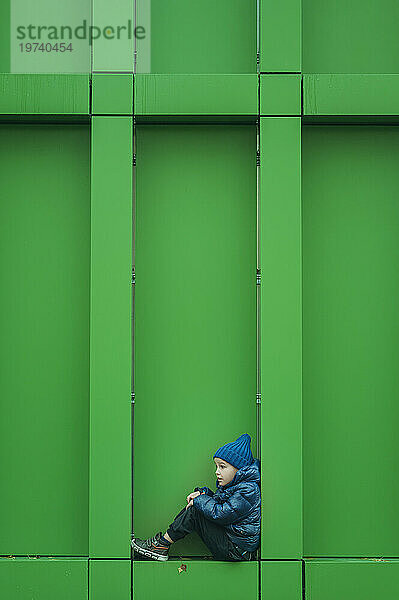 Boy in warm clothing sitting near green wall