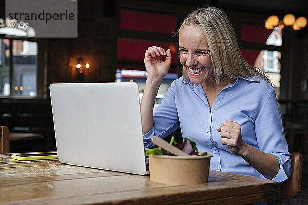 Glücklicher Freiberufler  der per Videoanruf über Laptop mit Salat auf dem Tisch in der Kneipe spricht