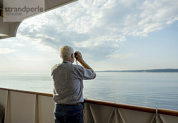 Älterer Mann blickt vom Schiff aus durch ein Fernglas auf das Meer