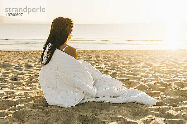 Frau sitzt in Decke gehüllt im Sand am Strand