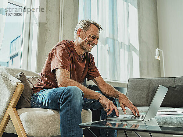 Glücklicher Mann  der zu Hause auf einem Sessel sitzt und einen Laptop benutzt