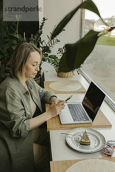 Blonder Freiberufler benutzt Smartphone und sitzt mit Laptop im Café