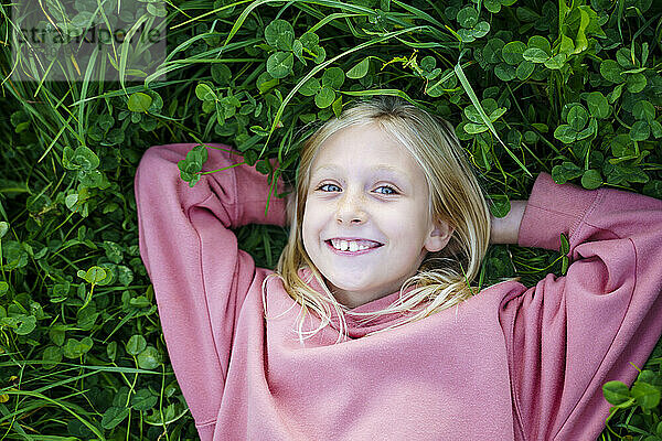 Lächelndes Mädchen in rosafarbenem Sweatshirt  die Hände hinter dem Kopf im Gras liegend