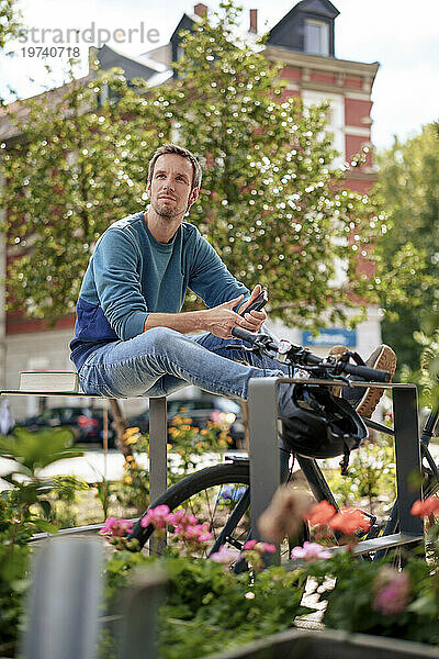 Glücklicher Mann hält Smartphone in der Hand und sitzt auf einer Metallkonstruktion neben dem Fahrrad