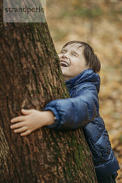 Fröhlicher Junge umarmt Baum im Herbstpark