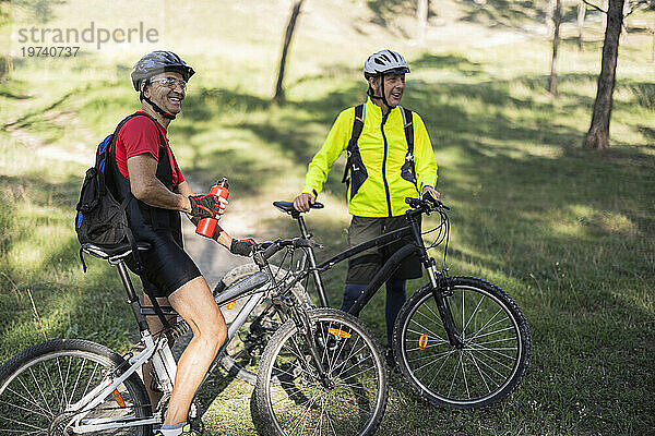 Lächelnder Mann hält Wasserflasche in der Hand und steht neben Freund mit Fahrrad im Wald