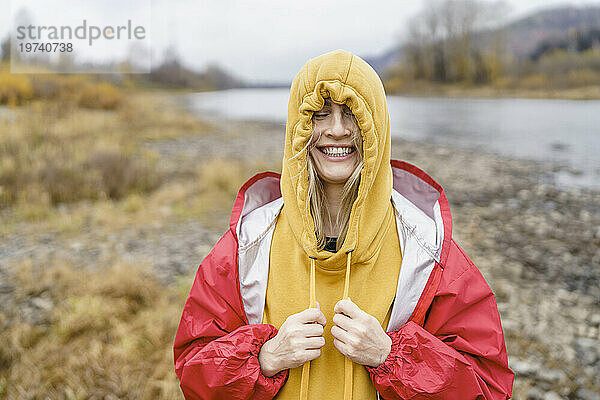 Fröhliche Frau zieht ihr Kapuzenshirt vor dem Fluss enger an