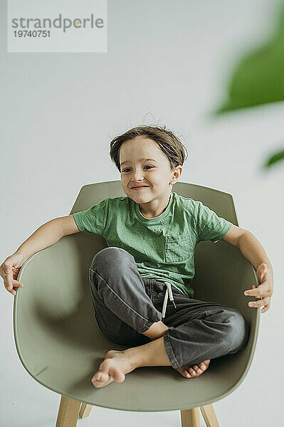 Glücklicher Junge sitzt auf einem Stuhl vor einer weißen Wand