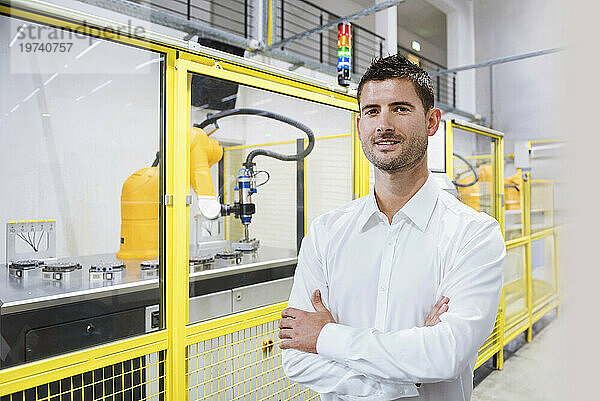 Lächelnder Ingenieur mit verschränkten Armen steht in der Fabrik