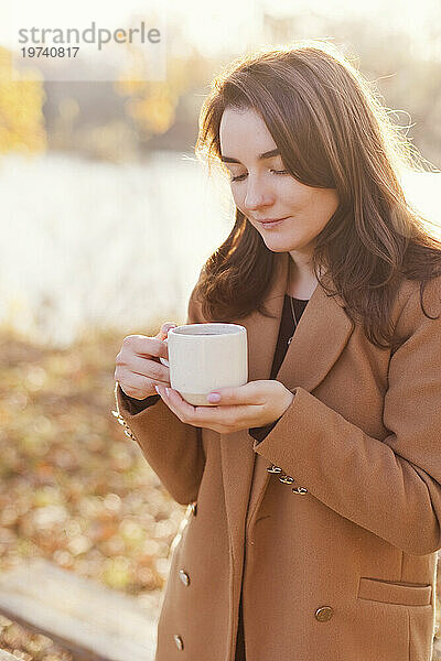 Glückliche Frau trägt Mantel und hält eine Tasse Tee im Herbstpark