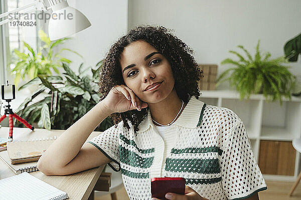 Smiling curly haired girl sitting with smart phone at desk
