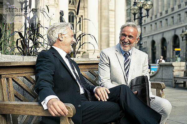 Fröhliche Geschäftsleute sitzen lachend auf einer Bank vor Finanzgebäuden