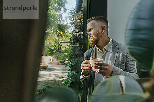 Smiling businessman looking through window and holding coffee cup in office
