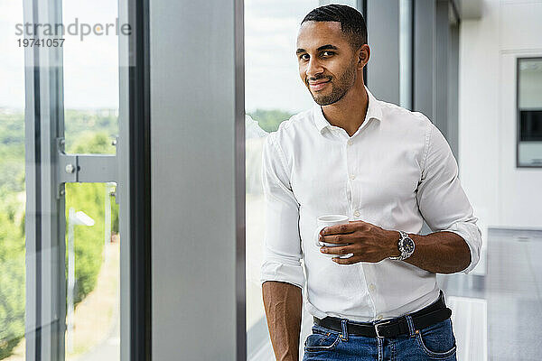 Casual businessman standing by window holding cup of coffee