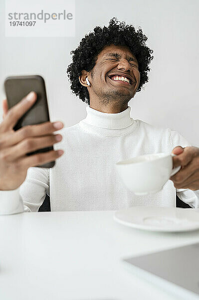Fröhlicher Freiberufler hält eine Tasse Tee und ein Smartphone am Schreibtisch