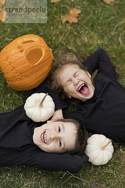 Excited siblings lying on grass with pumpkins