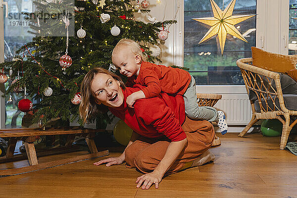 Lächelnder Sohn spielt zu Hause auf dem Rücken der Mutter neben Weihnachtsbäumen