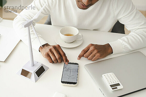 Geschäftsmann untersucht Windturbinendiagramm auf Smartphone am Schreibtisch im Heimbüro