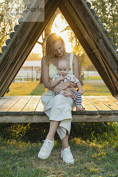 Liebevolle Mutter sitzt mit ihrem kleinen Jungen im dreieckigen Holzsitz bei Sonnenuntergang