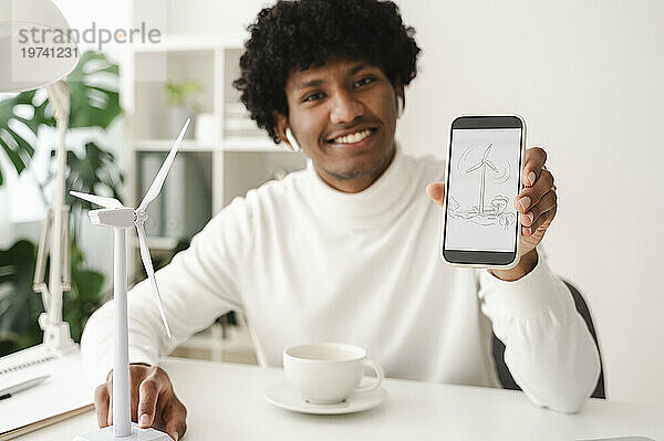 Lächelnder junger Geschäftsmann zeigt Diagramm einer Windkraftanlage auf Smartphone am Schreibtisch