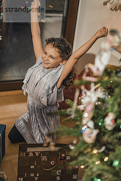 Smiling girl with arms raised kneeling near Christmas tree at home