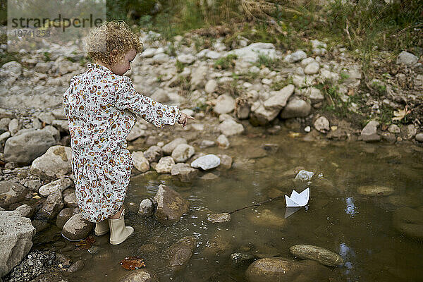 Nettes Mädchen spielt und zeigt auf ein Papierboot in einer Wasserpfütze in der Nähe von Felsen