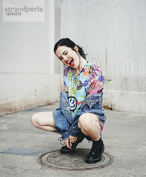 Young woman crouching on manhole and laughing in front of white wall