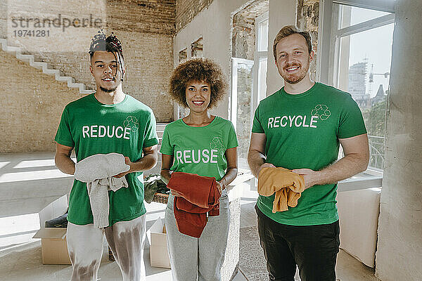 Lächelnde Aktivisten halten gebrauchte Kleidungsstücke in der Hand und stehen in der Wohnung