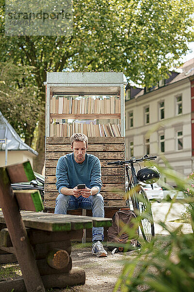Mann sitzt mit Smartphone auf Bank neben Bücherschrank