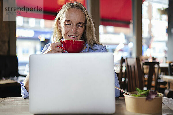 Lächelnder Freiberufler hält Kaffeetasse in der Hand und arbeitet am Laptop im Café