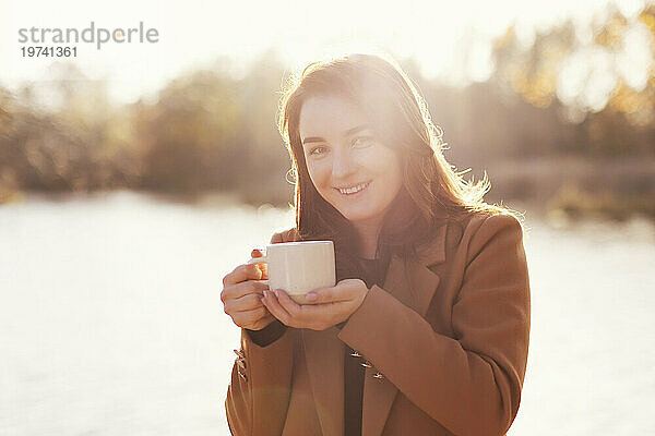 Lächelnde Frau hält bei Sonnenuntergang eine Tasse Tee am See