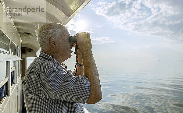 Neugieriger älterer Mann blickt vom Schiff aus durch ein Fernglas auf das Meer